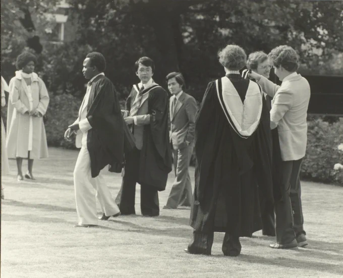 Black and white image of students from 1980 graduating, two men in centre of the picture look particularly stylish, with cuban heels and flares 