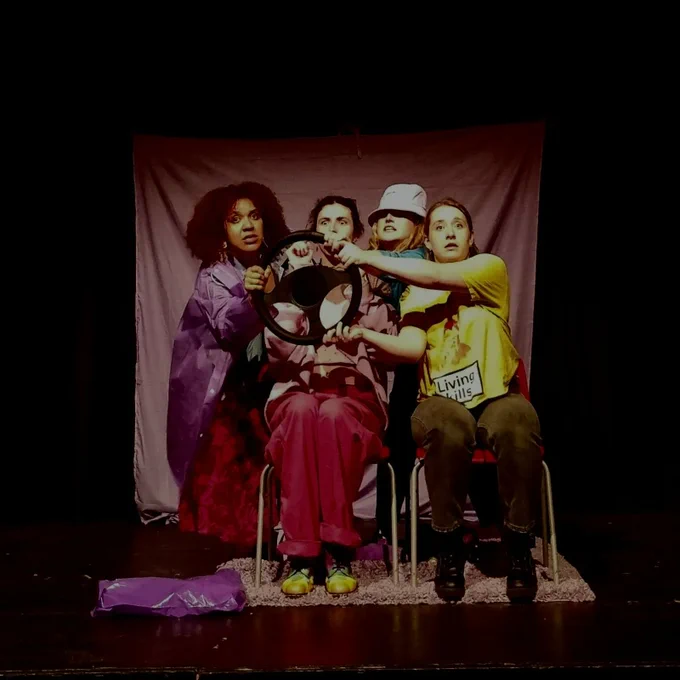 A production still from FOUR FELONS AND A FUNERAL, four people are sitting in chairs pretending to be car, holding a steering wheel and looking surprised