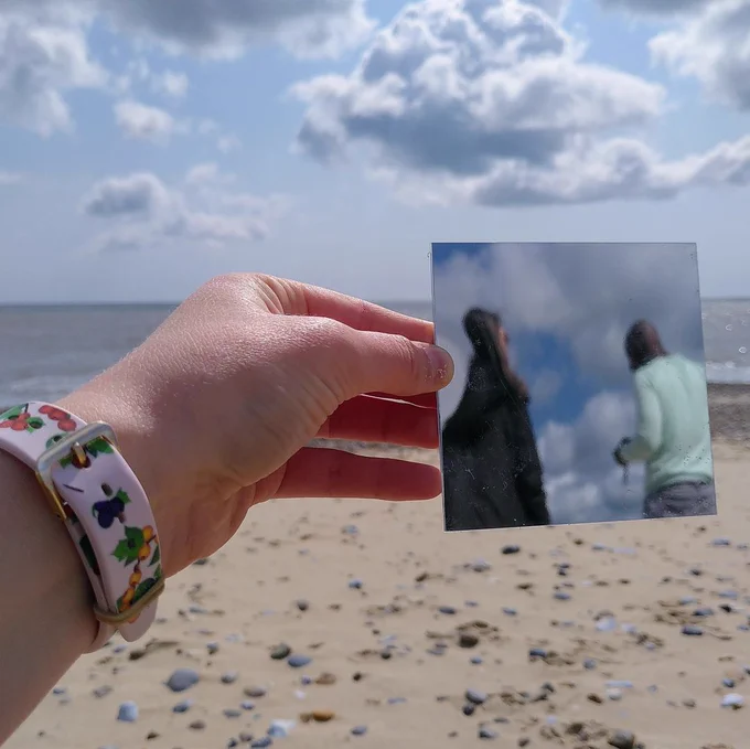 on a beach, a person wearing a pink floral watch holds out a square mirror from the left of the frame. In the mirror, a pair of people can be seen from behind