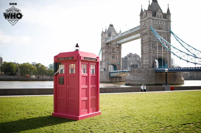 A photo of the TARDIS, painted bright pink, on the bank of the River Thames in London. The Tower Bridge can be seen behind it. A corner of the pink on the TARDIS appears to be peeling, revealing the original blue colour underneath.