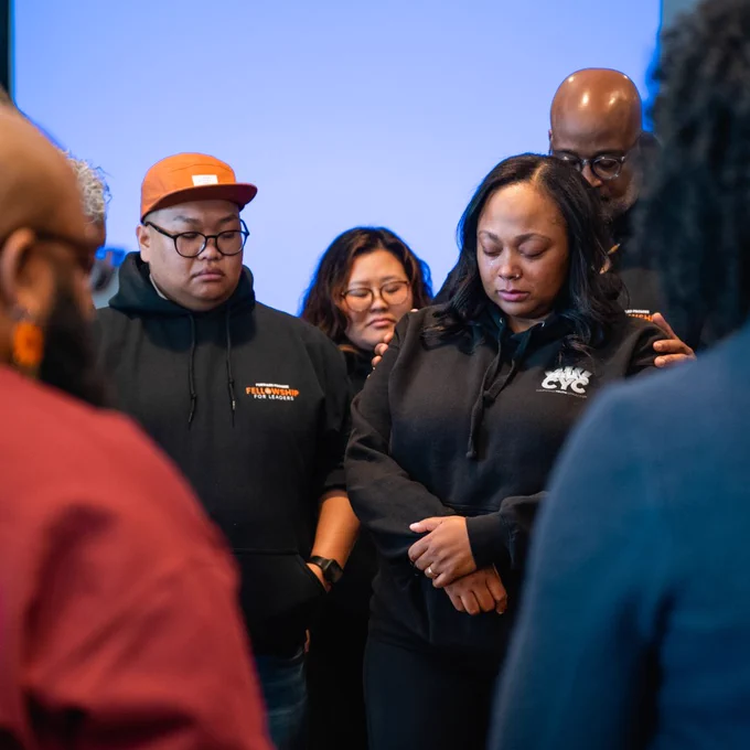 At March's fellowship retreat, fellows stand in a circle eyes closed. In the forefront is a Southeast Asian man wearing an orange baseball cap and black hoodie. Next to him is a Black woman with her head down and wearing a CYC black hoodie. A tall Black man stands behind her with hands on her shoulders.