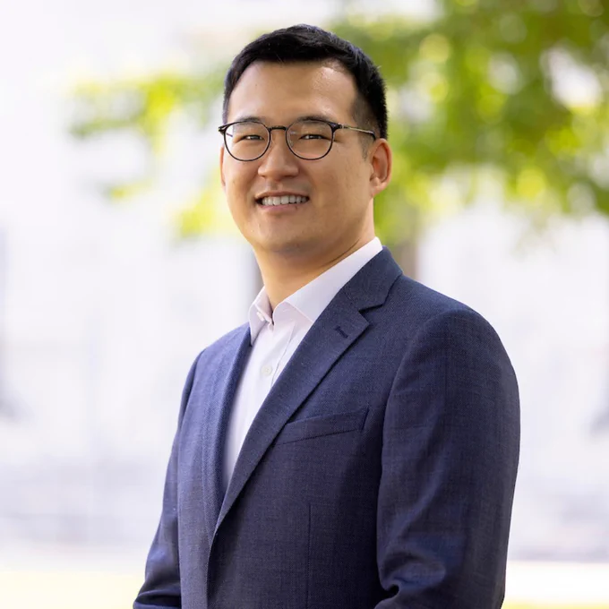 Chris Suh smiles outside of blurred trees and pink marble buildings on the Emory Quad. He wears a white button-down shirt and blue blazer