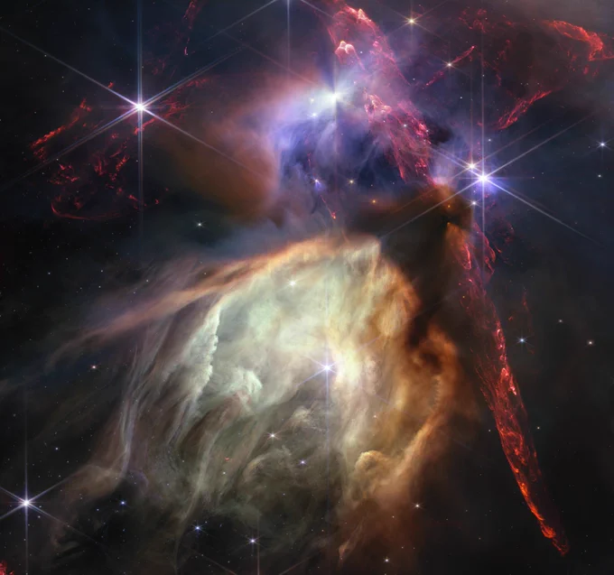 Dobles chorros rojos opuestos provenientes de estrellas jóvenes llenan la mitad superior más oscura de esta imagen, mientras que una estructura brillante de color amarillo pálido, similar a una cueva, está en el centro inferior, inclinada hacia las dos en punto, con una estrella brillante en su centro.