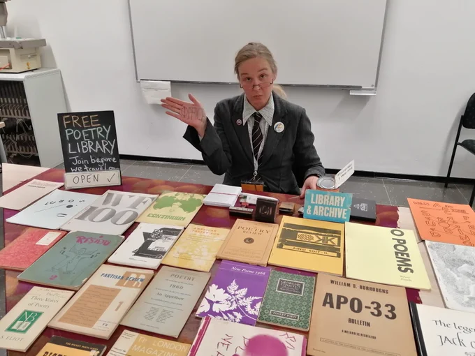 The Itinerant Poetry Librarian operating in Münster: The Librarian is sat in front of the Itinerant Poetry Library, with her "office" open, and that day's library selection of books on offer, including: W.S. Burroughs, Peter Finch, T.S. Eliot, Partisan Review, Bob Cobbing, "New Poems 1966" and Ciaran Carson.