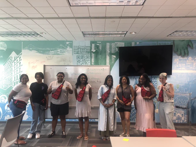 7 people stand side by side; they're wearing red cross body bags with stop the bleed supplies. The third person from the right is Roz Pichardo, trainer. 