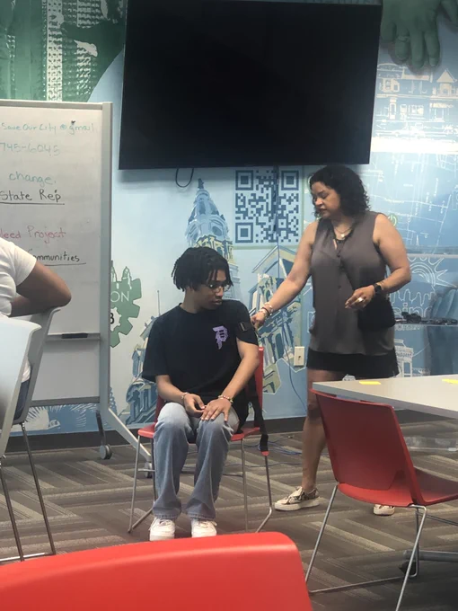 Roz Pichardo facilitates a training standing in the front of the room. A CRC member is serving as a demonstrative support, sitting in a chair next to Roz. 