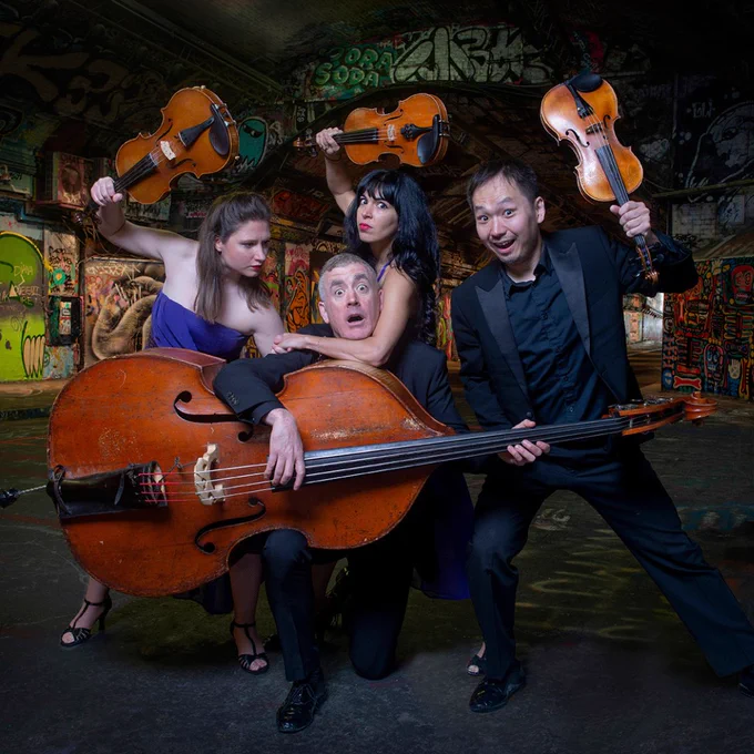 Two women and a man holding violins jokingly hold a man playing a cello. They're play fighting comically.