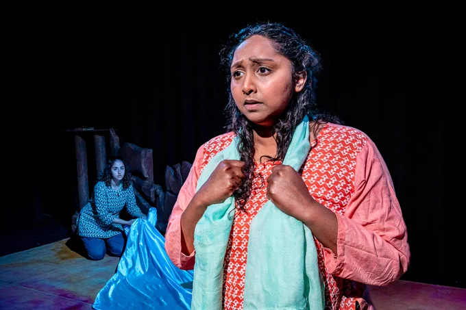 Two South Asian girls in traditional clothes, one in red/orange the other in greens. The one in orange has an aqua coloured dupatta around her shoulders; she looks sad and a little mystified. In the background the one in green is kneeling by a little wall, pulling in a large piece of shiny pale blue fabric