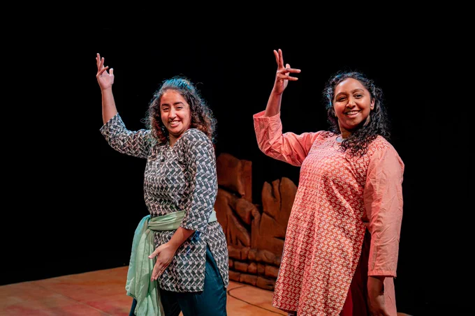 Two South Asian girls in traditional clothes, one in red/orange the other in greens. Both stand side by side, striking the same pose; with their right hands held up in a flourish gesture. They are smiling and laughing. 