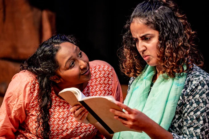 Two South Asian girls in traditional clothes, one in red/orange the other in greens. The one in orange is looking at her friend cheekily and though asking a question and giggling. The one in green is holding up a brown book as though she is reading it. She looks confused.