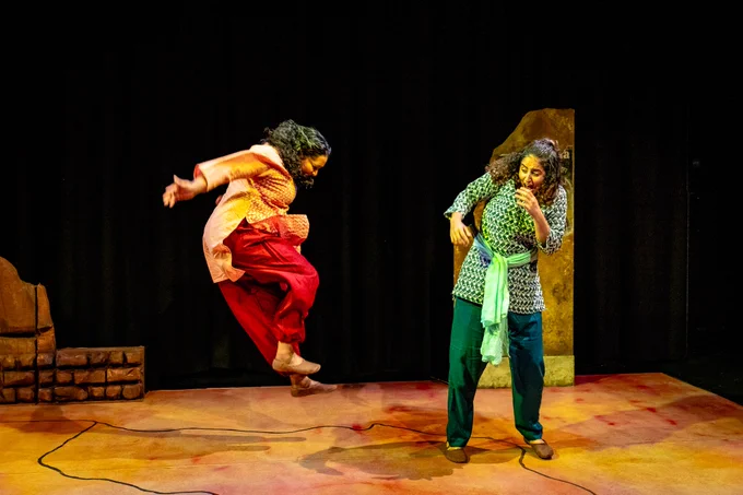 Two South Asian girls in traditional clothes, one in red/orange the other in greens. The one in red/orange is jumping forcefully in the air towards her friend. The one in green is screaming with laughter at her friend. The are in a theatre on a set made up of an orange, yellow and pinkish floor and there are two pieces of wall at the back. 