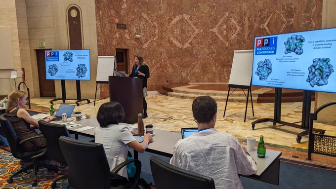 Jeanine Amacher presents research from a podium in front of a coral-colored granite wall at a hall at the Bolger Center in Potomac, Maryland.