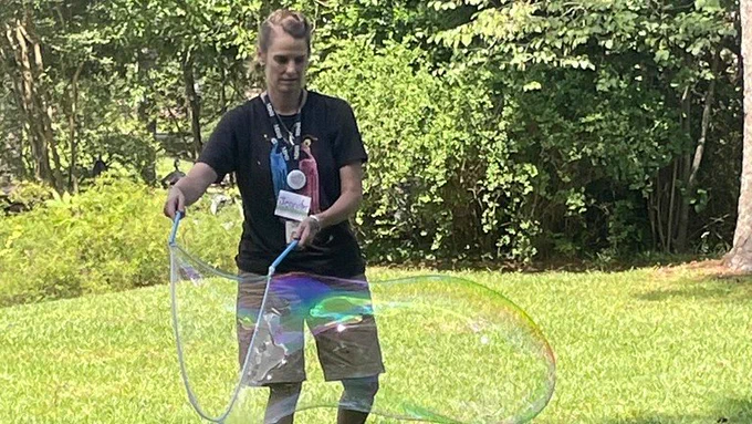 Jennifer Rose, a white woman, makes a large soap bubble via a bubble wand.
