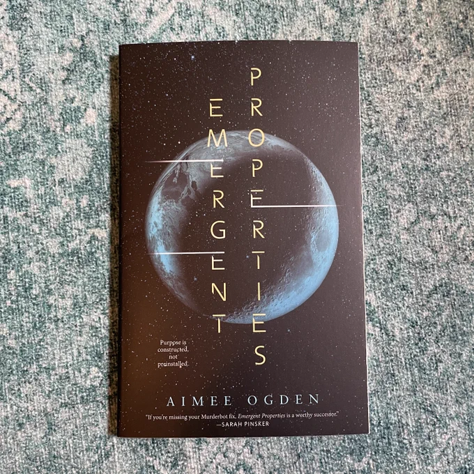 A copy of Emergent Properties by Aimee Ogden resting on a teal, patterned fabric background.