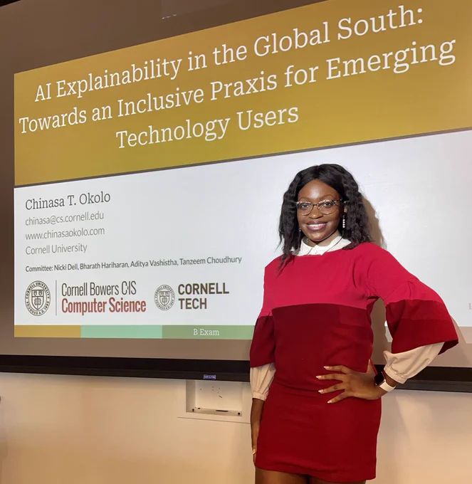 Chinasa is wearing a red dress with a white shirt underneath and is smiling.  Behind her is the title slide from her thesis defense “AI Explainability in the Global South: Towards an Inclusive Praxis for Emerging Technology Users”