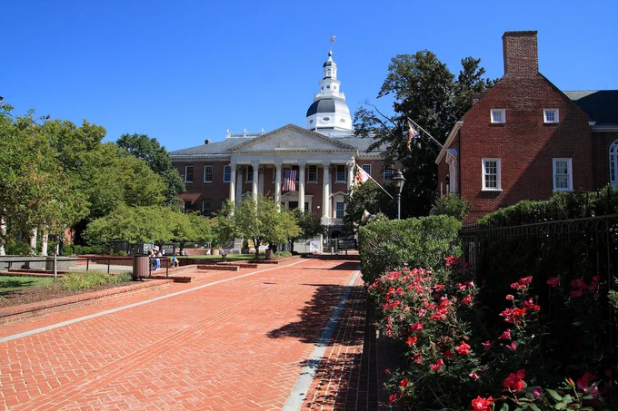 Maryland State House, with red and white elements, before blue sky and behind green trees and red houses