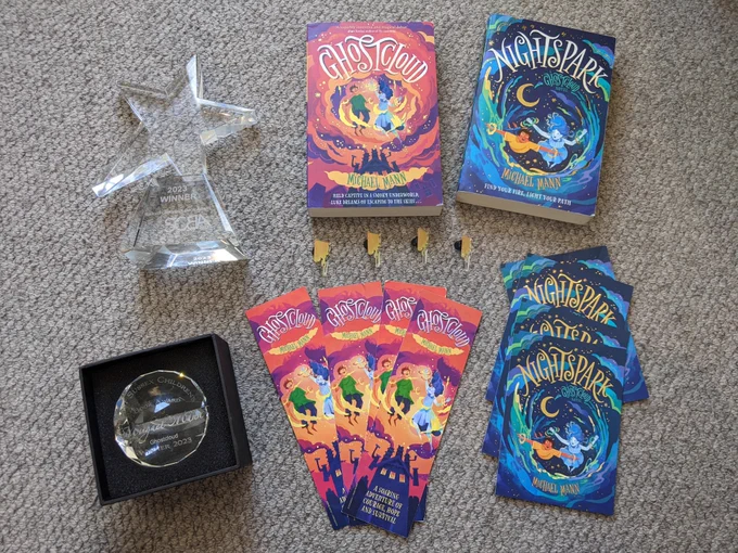 Pin badges, trophies, bookmarks, and a copy of the books Ghostcloud and Nightspark by Michael MAnn