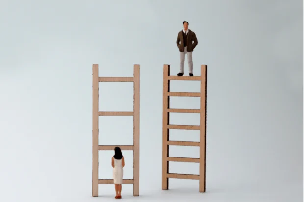 The miniature figure of a woman stands on the ground facing a wooden ladder. Beside her, a miniature male figurine stands atop another wooden ladder.