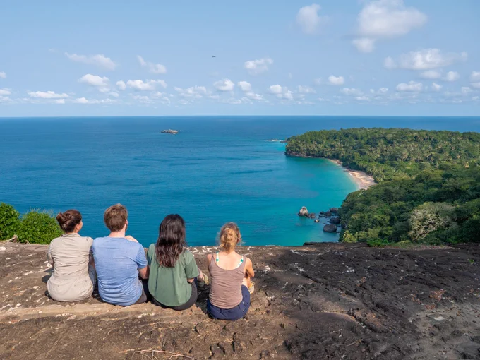 The expedition team are sitting on a cliff overlooking a view of the ocean and rainforest. 