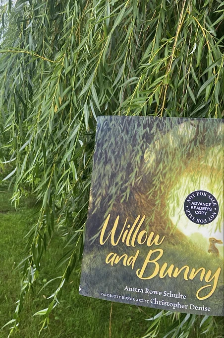 Photograph is a picture book (advanced reader copy) held out in front of green, swaying willow tree branches. The book is WILLOW AND BUNNY, which features a broad willow tree on the left and a small bunny on the right. The title is in sunny yellow script on the bottom half of the book.