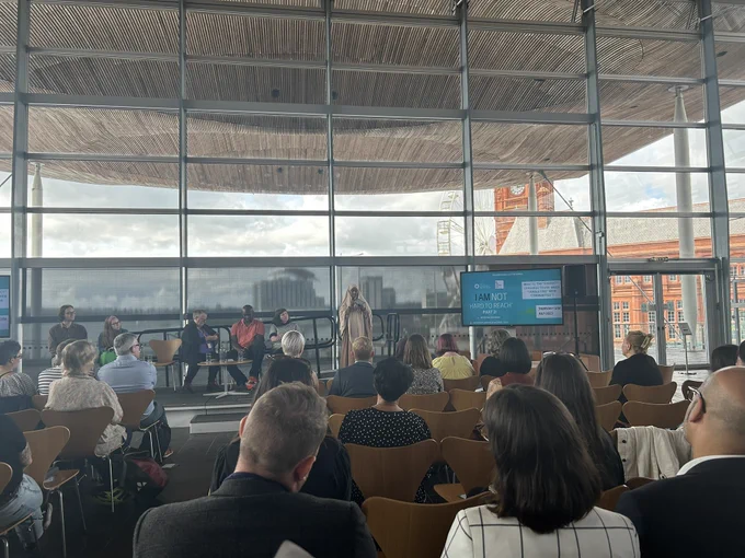 Image from an event in the Senedd. Speakers on the stage in front of an audience. Cardiff Bay in the background.
