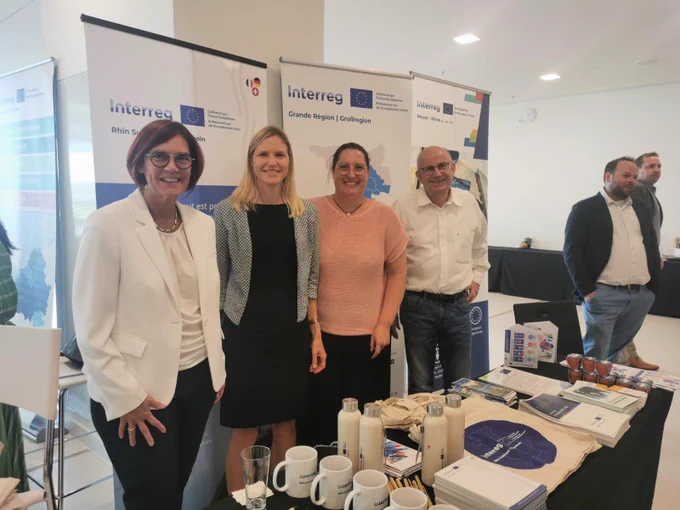 Im Hintergrund drei Roll-ups der Interreg A Programme. Davor stehen vier Personen: von links nach rechts Staatssekretärin Petra Dick-Walter, Katarina Klein (SGD Süd für Oberrhein), Claudia Krütten (ADD für Maas-Rhein) und Dieter Müller (ADD für Großregion). Davor steht ein länglicher Tisch, auf dem Infomaterial sowie give-aways wie Becher, Trinkflaschen, Stoffbeutel, Blumentöpfe mit Samen liegen.