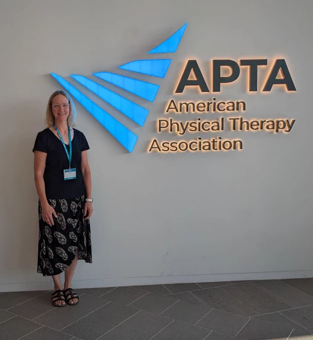 Image shows Dr. Laura Gras smiling next to a wall with the logo for the American Physical Therapy Association