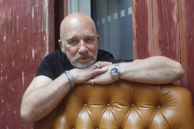 Headshot of architect Nigel Coates, smiling with his arms folded resting on a light brown armchair. He wears a black t-shirt, a watch, bracelets, and two rings.