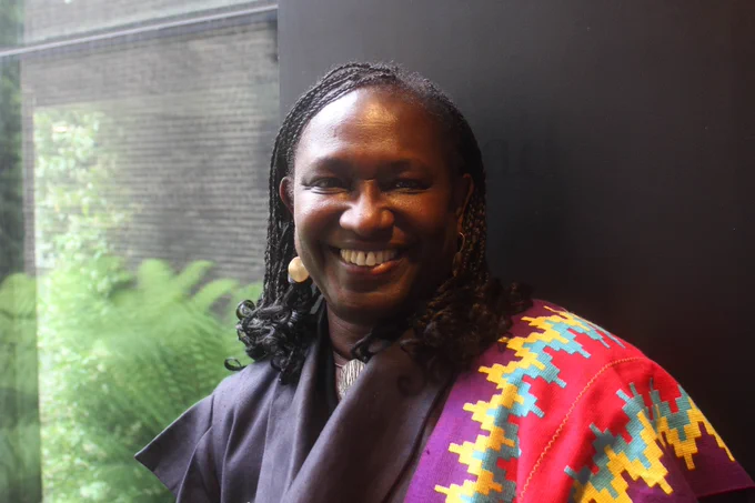 Headshot of architect Elsie Owusu, smiling in front of a window and a grey wall. The window overlooks a building and a green plant.