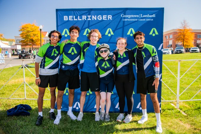 A group of six BellRinger participants pose for a group photo in front of a dark blue and green BellRinger backdrop