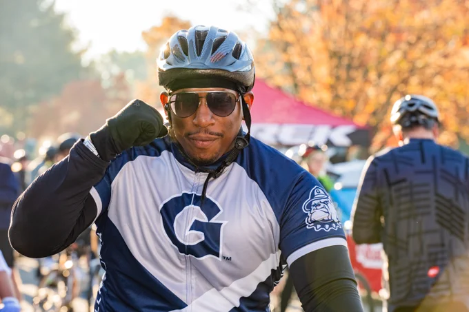 A cyclist on their bike makes a fist with their right hand and smiles at the camera. The cyclist is wearing a Georgetown cycling jersey, and has a helmet and sunglasses on.