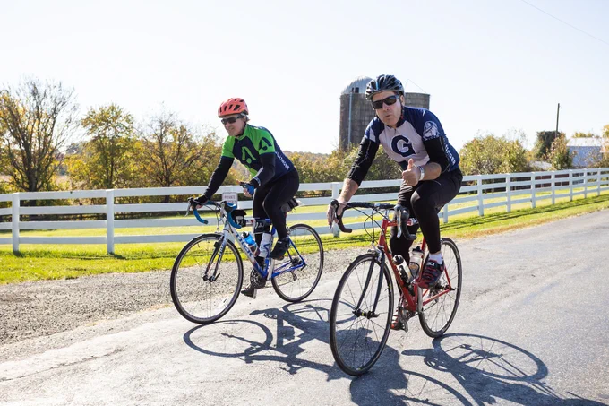 Two cyclists bike down a road lined with a white fence. The cyclist closest to the camera gives a "thumbs up."
