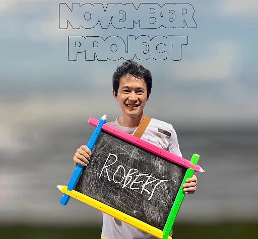 A waist-up yearbook photo picture of Robert Ling who is holding a chalk sign with his name on it. November Project logo is overlaid above his head. 