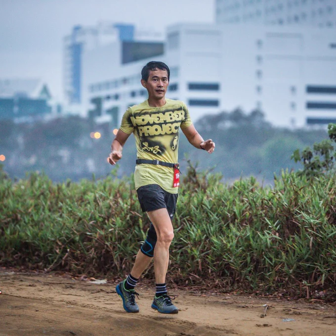 A photo of Robert Ling participating in the trail race. He is running on a dirt road that is lined with waist-deep bushes and shrubs wearing black shorts and a yellow t-shirt with November Project tag on it. Some buildings are off-focus but visible in the background. 