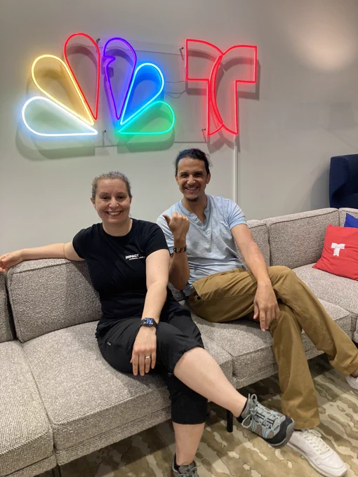 Meg & Josh sitting on a couch smiling with the lit-up NBC logo behind them