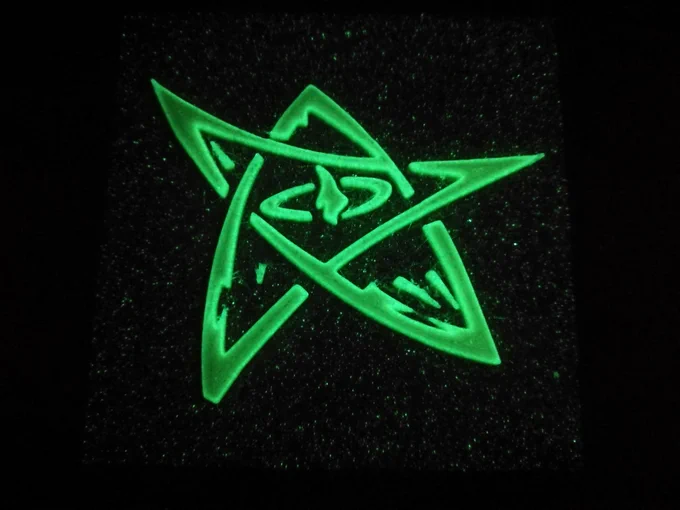 A 3D printed Elder Sign from the Cthulhu Mythos, glowing in the dark in green, over a background with tiny star-like green glowing dors. The outline of the Sign glows brighter than its inside.