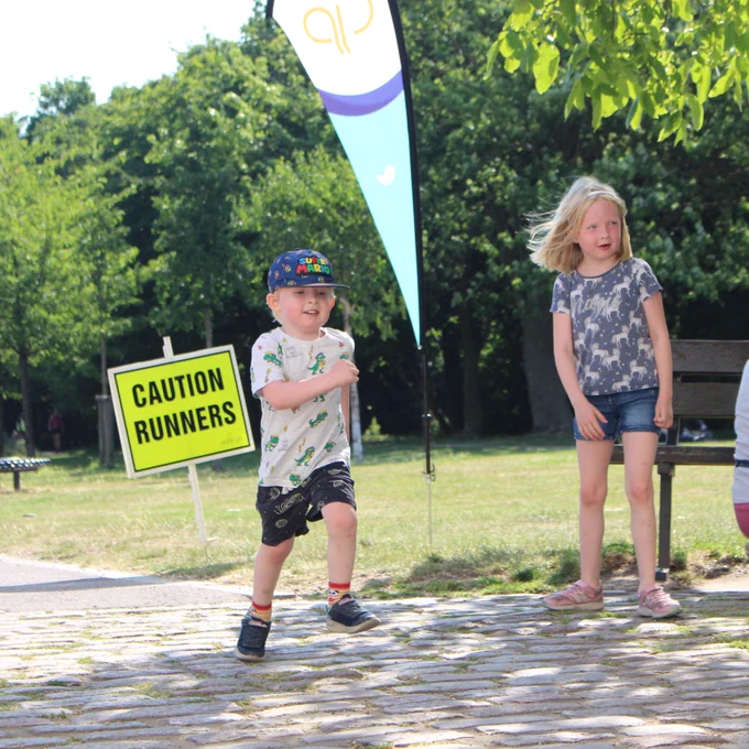 Child jogging in park, with parkrun flag and 'caution runners' behind.