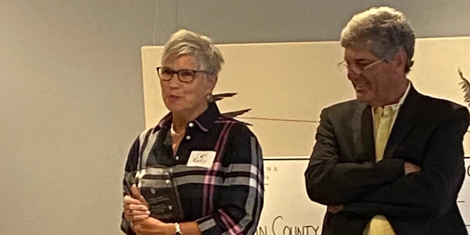Marty Driver, at left, speaks at podium holding her award. Mark Diachenko, standing to her right, looks on.