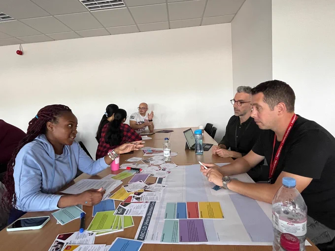 Photo of colleagues in small groups, sitting at desks talking and collaborating with cards, paper and pens 