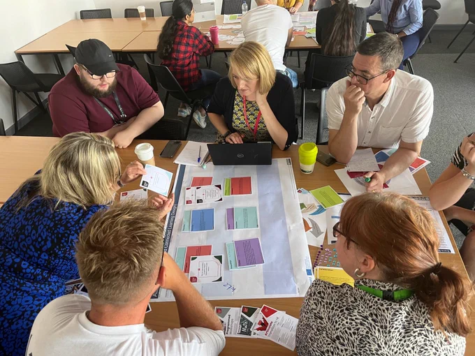 Photo of colleagues in small groups, sitting at desks talking and collaborating with cards, paper and pens 
