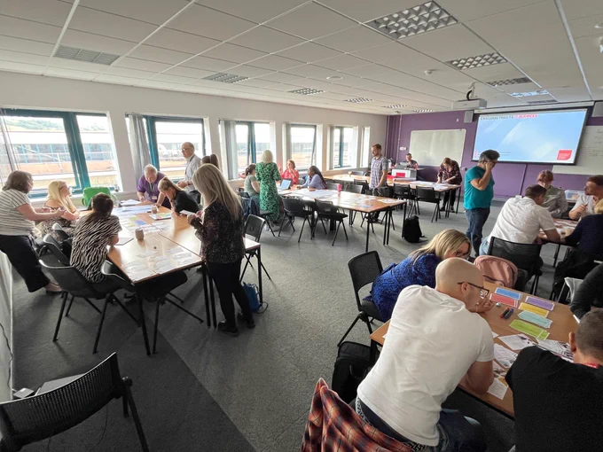 Photo of colleagues in small groups, sitting at desks talking and collaborating with cards, paper and pens 