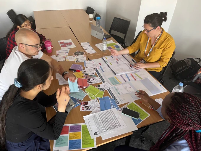 Photo of colleagues in small groups, sitting at desks talking and collaborating with cards, paper and pens 