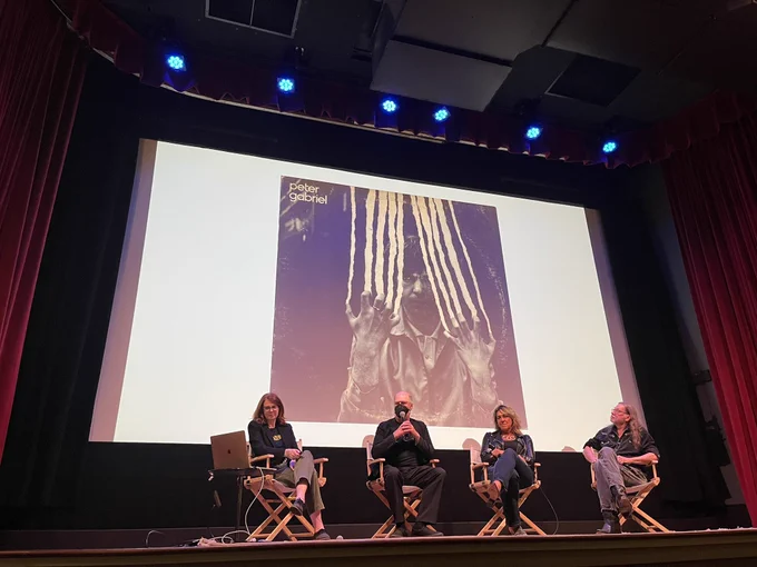 Four people on a stage with a Peter Gabriel album cover, designed by Hipgnosis, in the background.