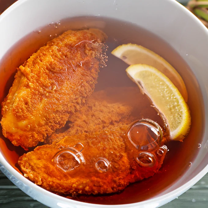 Zwei panierte Schnitzel in einer weißen Schüssel versinken in einer roten Flüssigkeit, zu ihnen gesellen sich zwei Stücken Zitrone. Aus irgendeinem Grund blubbert das Ganze.