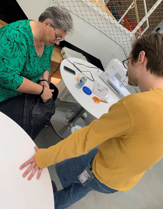 A woman holds a pair of pants that need a pocket repair while a man looks on. A sewing machine is on a table behind them.