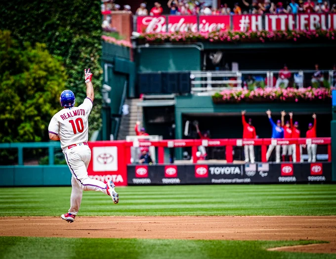 Photo of J.T. Realmuto rounding second base and giving the bullpen a peace sign with his right hand after hitting a home run.  He is wearing the white Phillies uniform with a blue and red helmet.