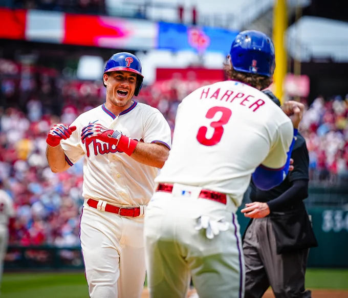 Photo of J.T. Realmuto smiling at Bryce Harper after hitting a home run.  They are both wearing white Phillies uniforms with blue and red helmets.