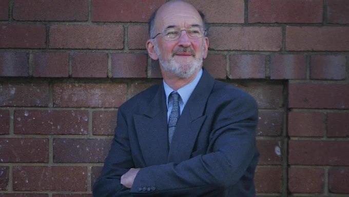 Photo of Hugh Saddler standing with arms folder and looking to the side in front of a brick wall. Hugh is wearing a dark blue suit.