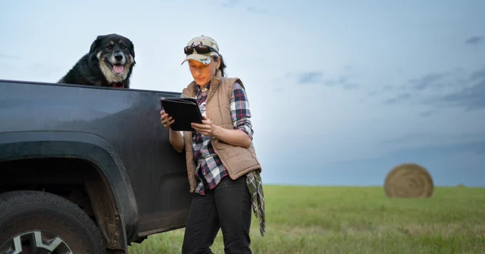 Woman holding tablet in a field. A dog is in the car behind her. 