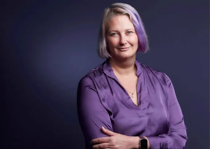 Portrait photo of Anna, who is wearing a bright purple silk shirt, that matches purple streaks in her light ash blond hair. She is smiling. She is set against a dark navy gradient background. 